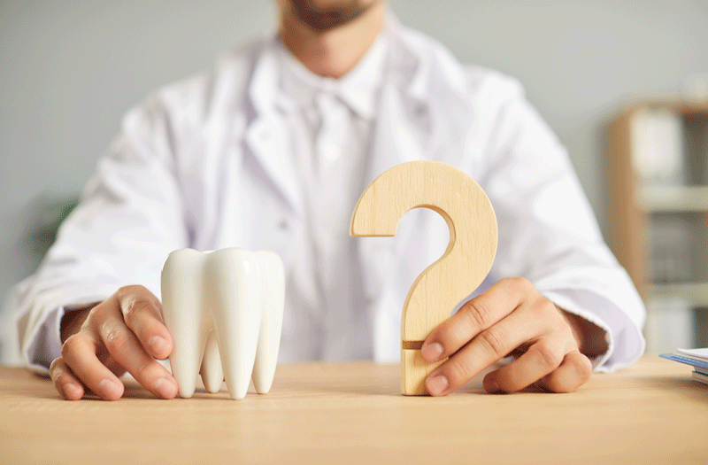 Professional dentist ready to answer all your questions about teeth health. Closeup male hands holding a white tooth model and a wooden question mark on the table.